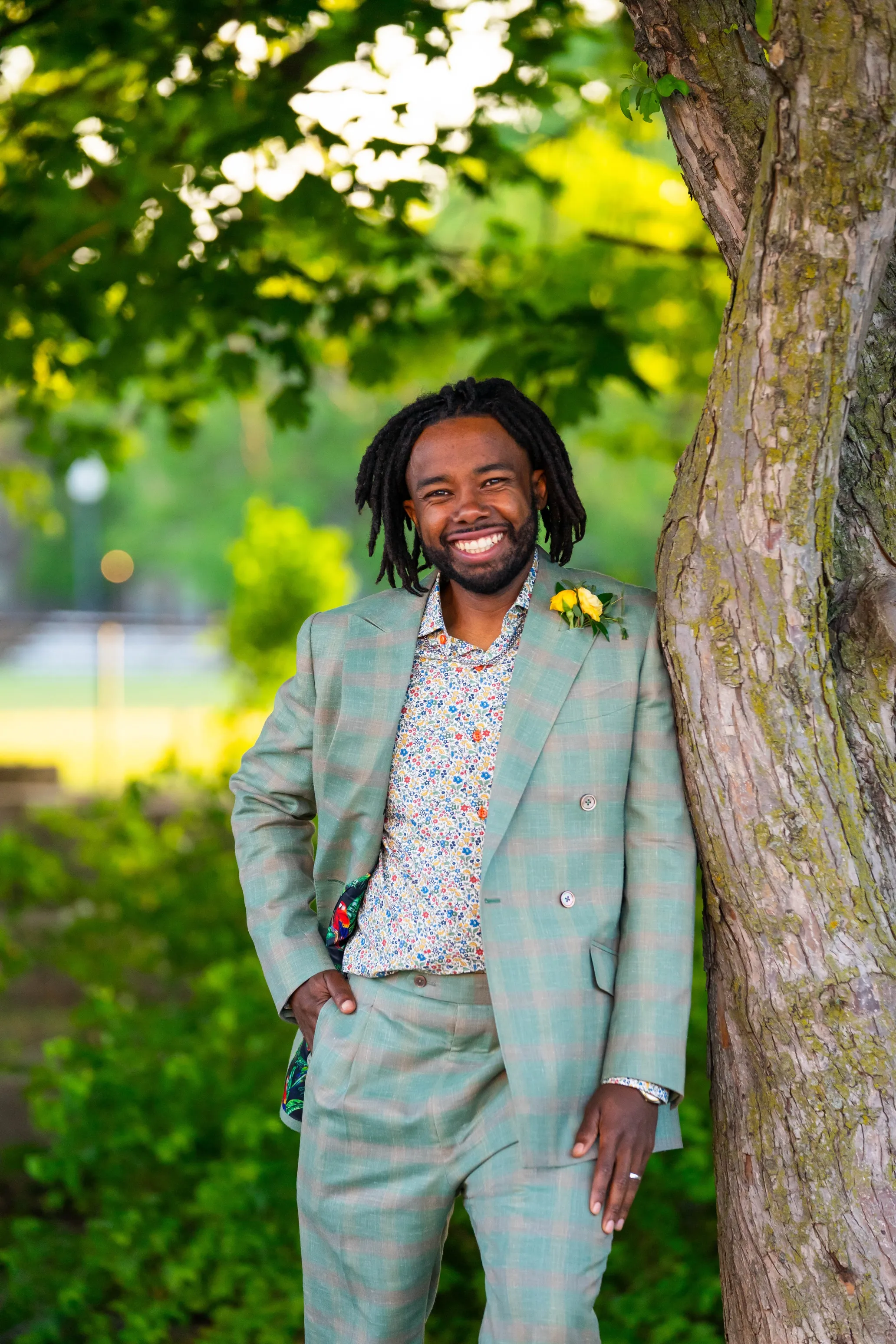 Anwar Eaton smiling outdoors, leaning against a tree in a green plaid blazer and floral shirt.